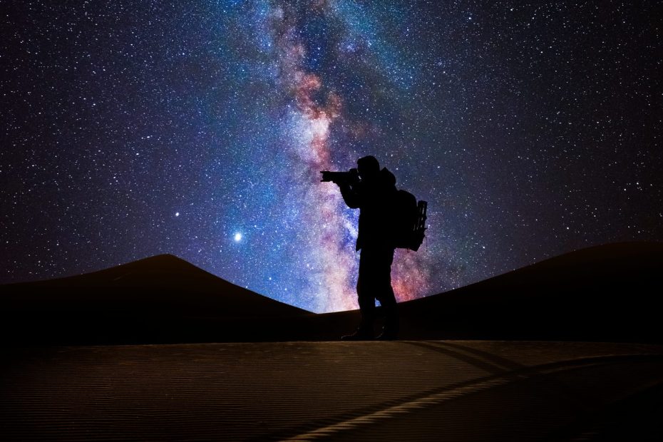 Photographe installé sur un trépied sous la Voie lactée, dans un paysage nocturne.